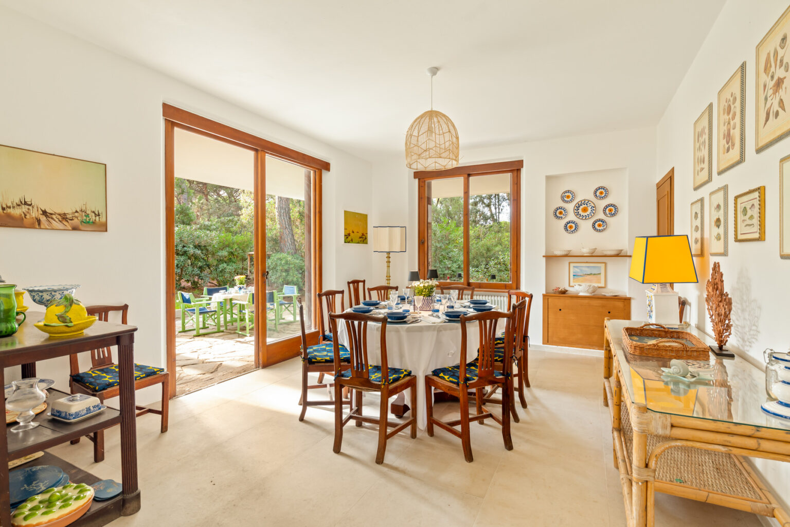 Bright and airy dining room featuring a round table set for six, surrounded by wooden chairs with vibrant blue cushions. The room has large glass doors opening to a lush garden, showcasing outdoor seating. Decorated with framed botanical prints and colorful plates, the space exudes a cozy, inviting ambiance. A stylish lamp and decorative items enhance the decor. | Only on SopranoVillas