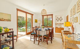 Bright and airy dining room featuring a round table set for six, surrounded by wooden chairs with vibrant blue cushions. The room has large glass doors opening to a lush garden, showcasing outdoor seating. Decorated with framed botanical prints and colorful plates, the space exudes a cozy, inviting ambiance. A stylish lamp and decorative items enhance the decor.