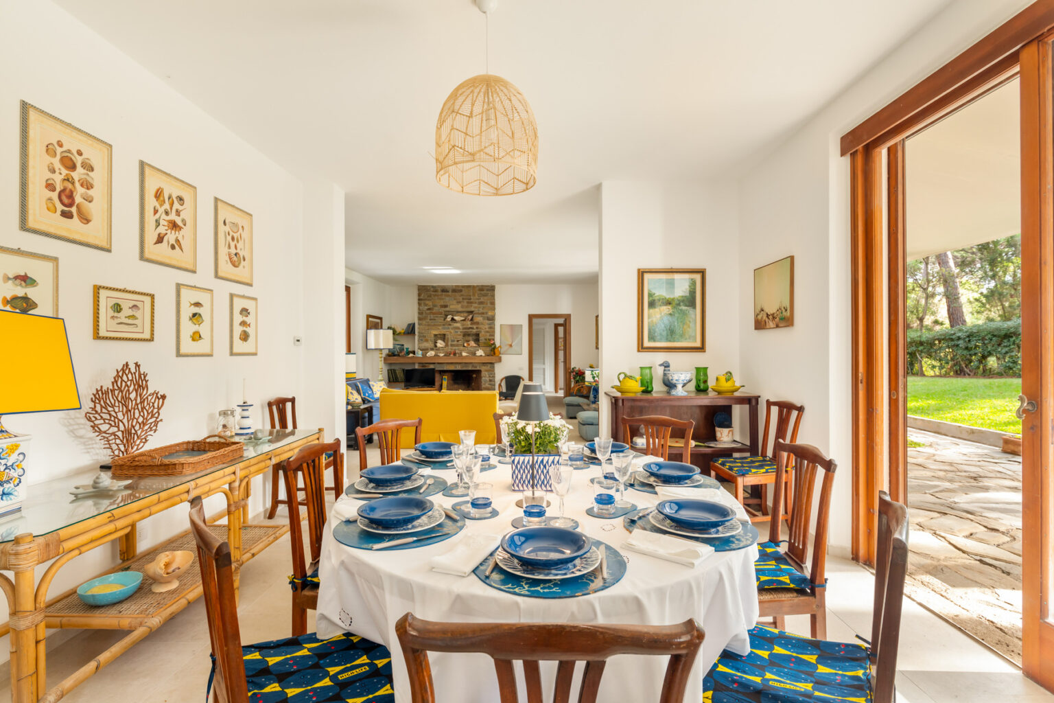 Bright and inviting dining area featuring a round table set with blue dishware, surrounded by wooden chairs. The room is decorated with framed botanical prints and a stylish lamp, with large windows offering a view of the garden. | Only on SopranoVillas