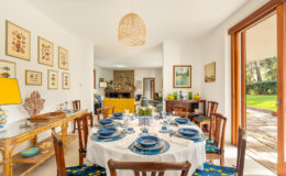 Bright and inviting dining area featuring a round table set with blue dishware, surrounded by wooden chairs. The room is decorated with framed botanical prints and a stylish lamp, with large windows offering a view of the garden.