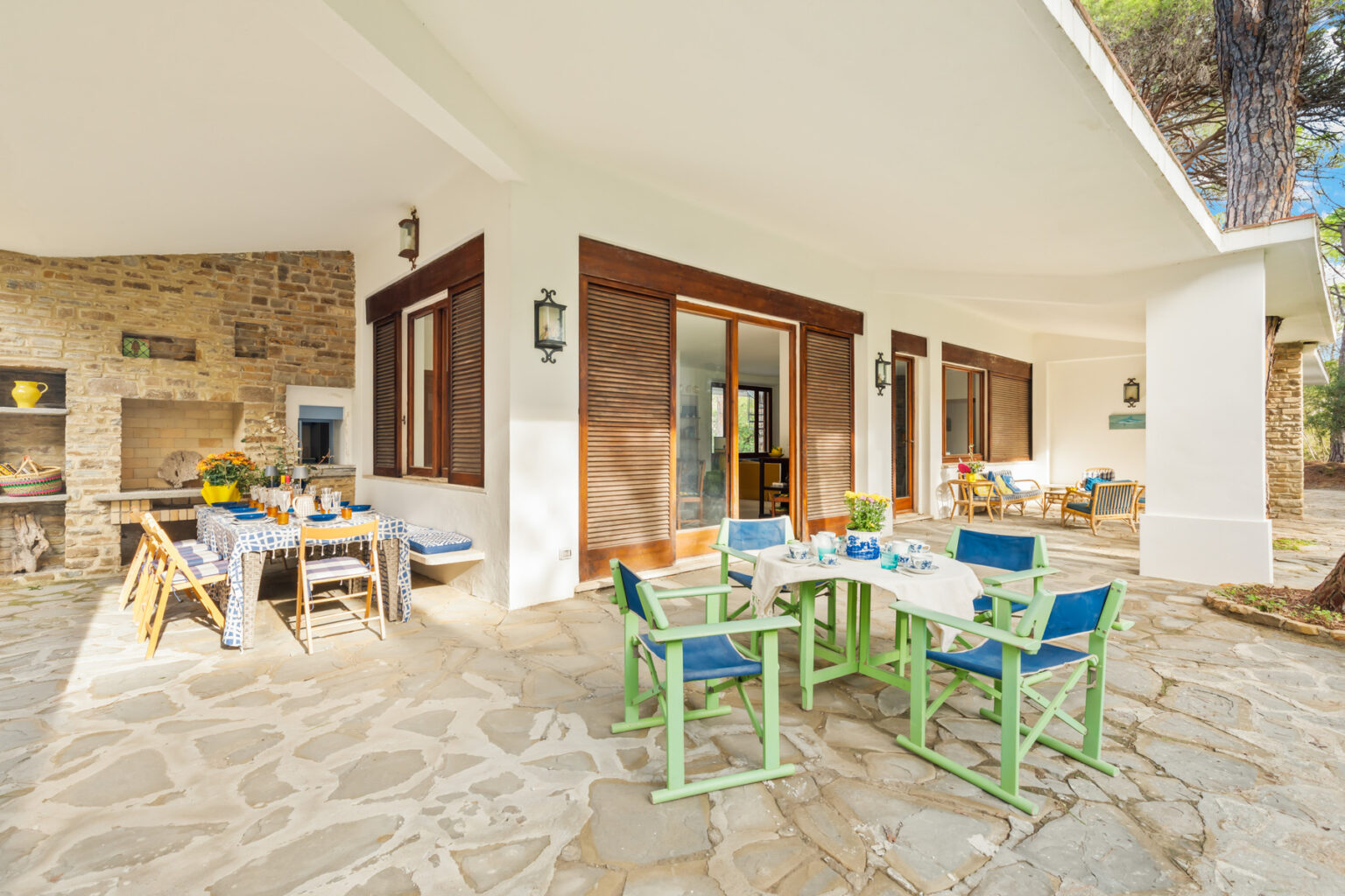 Outdoor dining area featuring a stone patio, colorful chairs, and a table set for a meal, surrounded by a modern home with large windows and a rustic stone wall. Ideal for relaxation and entertaining in a serene setting. | Only on SopranoVillas