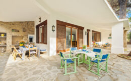 Outdoor dining area featuring a stone patio, colorful chairs, and a table set for a meal, surrounded by a modern home with large windows and a rustic stone wall. Ideal for relaxation and entertaining in a serene setting.