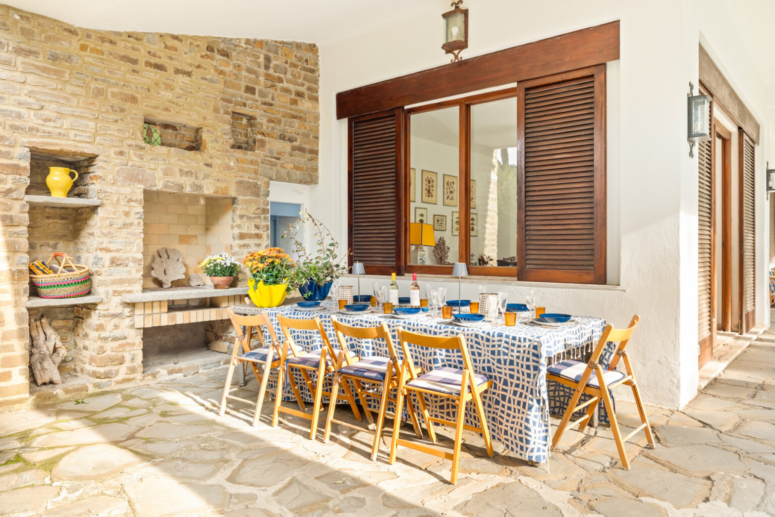 Outdoor dining area featuring a long wooden table set with blue dishes and yellow accents, surrounded by wooden chairs. The space includes a stone fireplace and large windows, creating a cozy and inviting atmosphere for gatherings. | Only on SopranoVillas