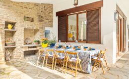 Outdoor dining area featuring a long wooden table set with blue dishes and yellow accents, surrounded by wooden chairs. The space includes a stone fireplace and large windows, creating a cozy and inviting atmosphere for gatherings.