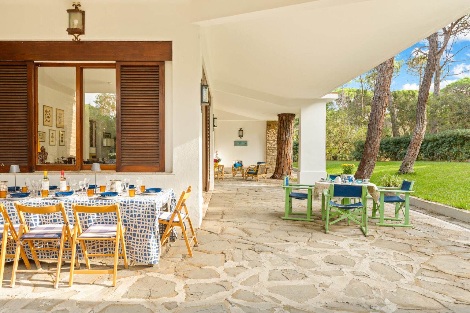 Outdoor dining area with a stone floor, featuring a large table set with colorful tableware and surrounded by wooden chairs. The space is illuminated by natural light and framed by lush greenery and trees, creating a serene atmosphere for gatherings. | Only on SopranoVillas