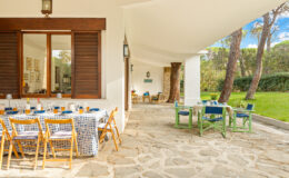 Outdoor dining area with a stone floor, featuring a large table set with colorful tableware and surrounded by wooden chairs. The space is illuminated by natural light and framed by lush greenery and trees, creating a serene atmosphere for gatherings.