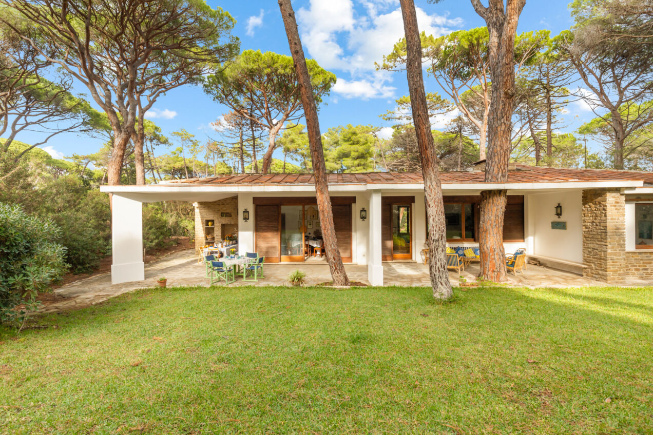 Modern single-story house surrounded by tall pine trees and lush green grass, featuring a spacious outdoor area with seating and large windows for natural light. | Only on SopranoVillas