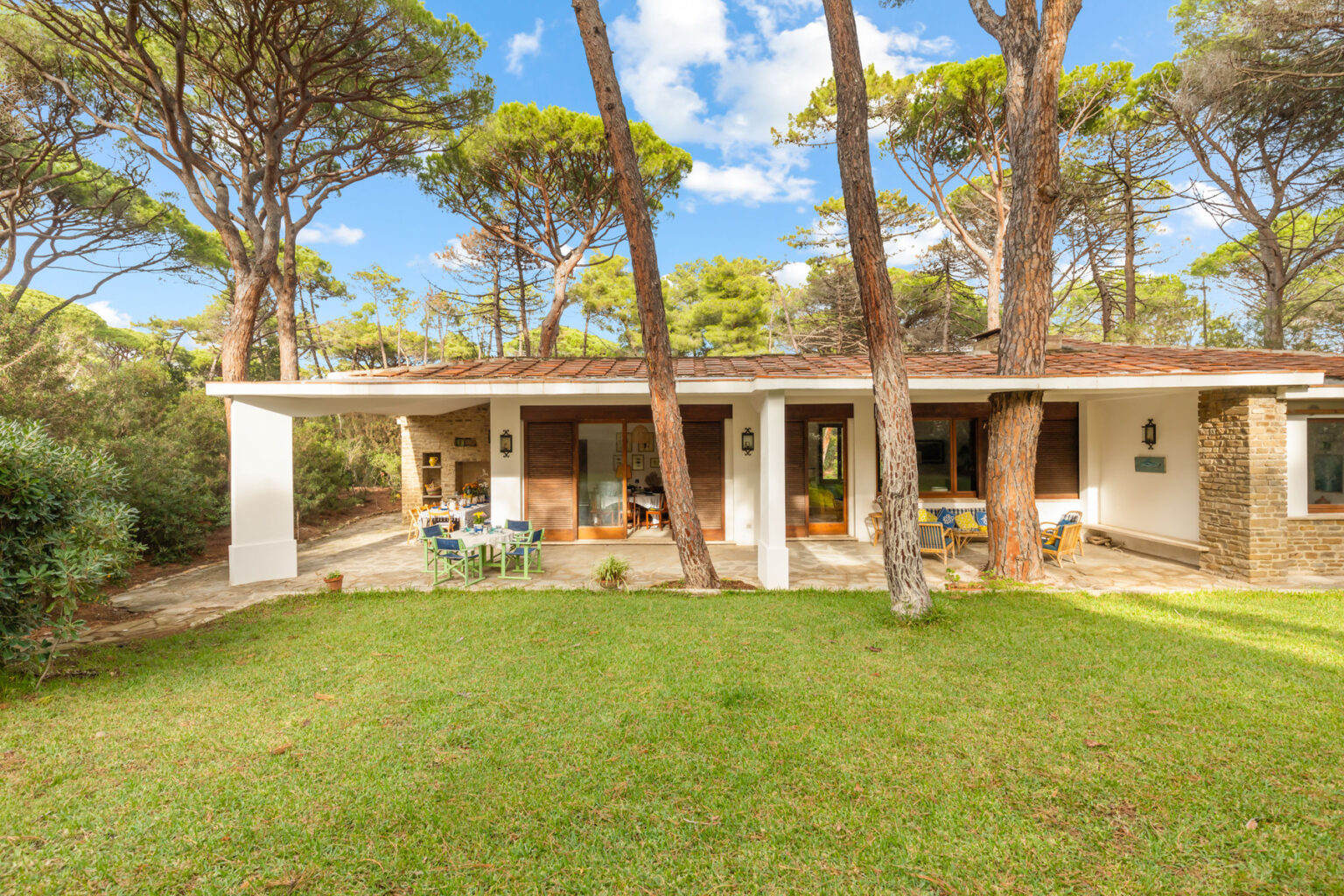 Modern single-story house surrounded by tall pine trees and lush green grass, featuring a spacious outdoor area with seating and large windows for natural light. | Only on SopranoVillas