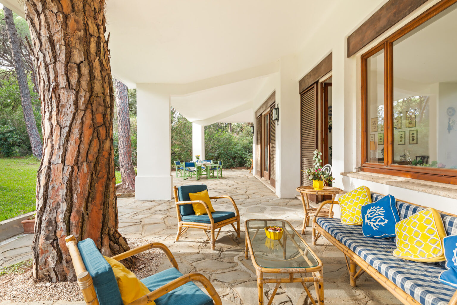 Cozy outdoor patio area featuring comfortable seating, a glass coffee table, and vibrant decorative pillows, surrounded by lush greenery and large windows that invite natural light. | Only on SopranoVillas