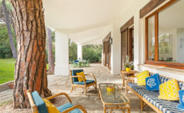 Cozy outdoor patio area featuring comfortable seating, a glass coffee table, and vibrant decorative pillows, surrounded by lush greenery and large windows that invite natural light.