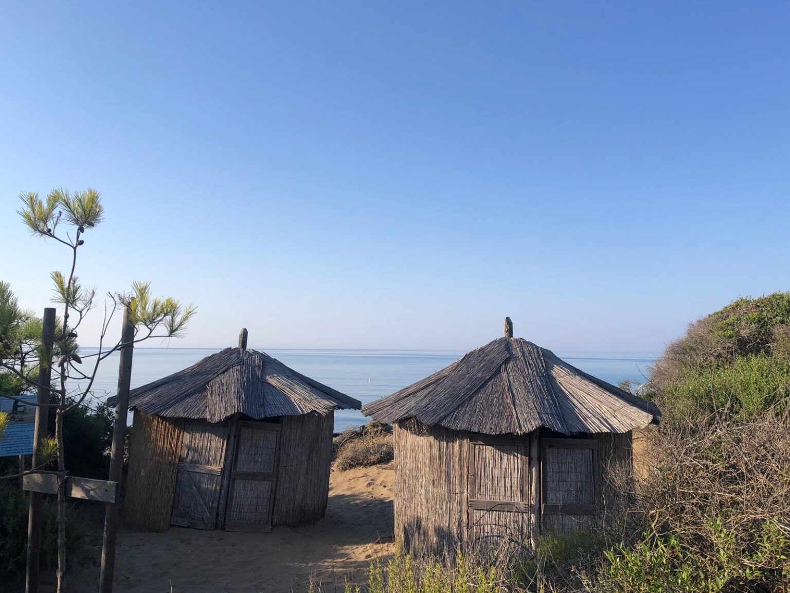 Two rustic beach huts surrounded by greenery, overlooking a calm sea under a clear blue sky. Ideal for a serene coastal getaway. | Only on SopranoVillas