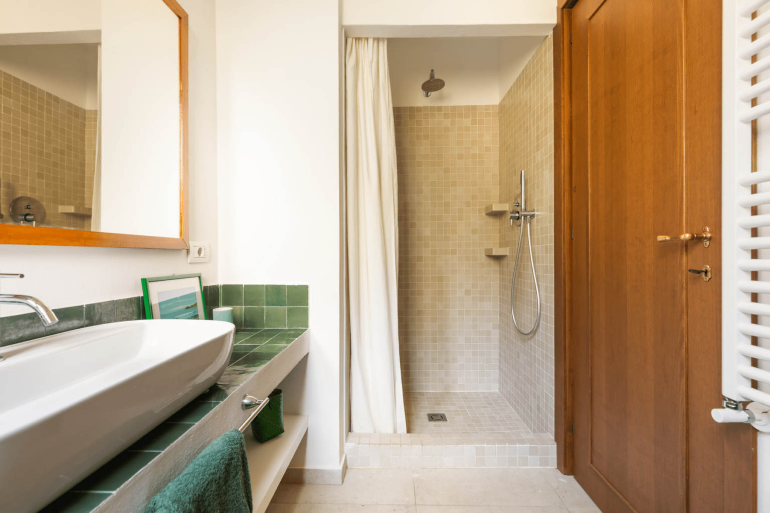 Modern bathroom interior featuring a sleek sink with green accents, a spacious shower area with a glass door, and wooden cabinetry. Natural light enhances the clean design and neutral color palette. | Only on SopranoVillas