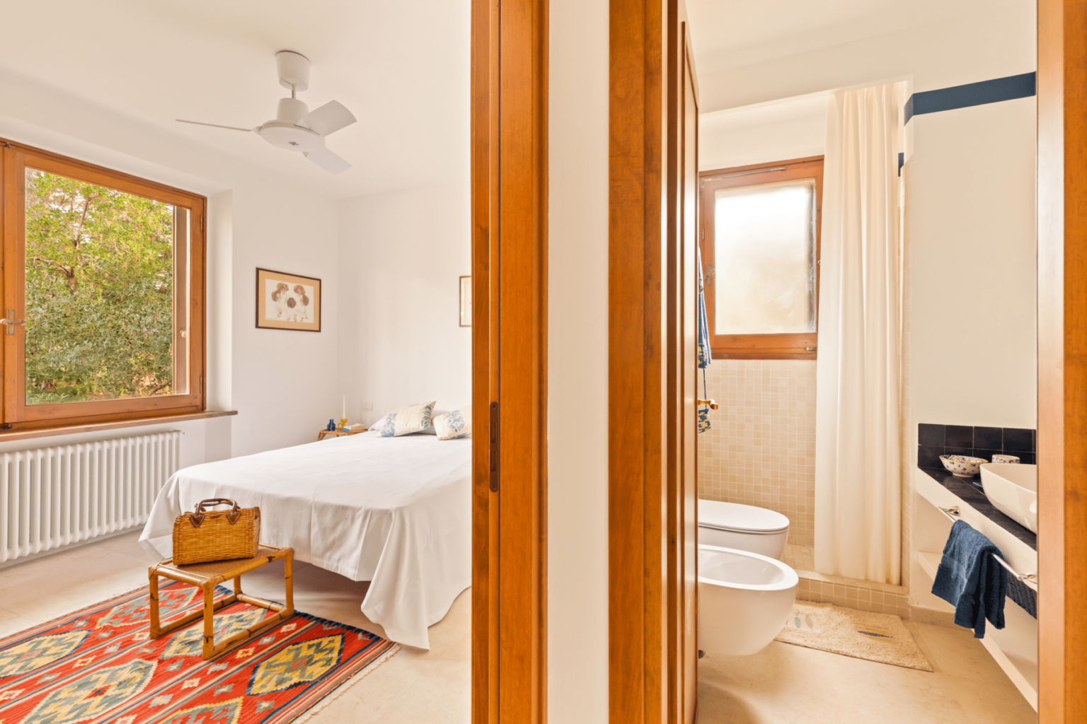Bright and airy bedroom featuring a white bedspread, wooden furniture, and large windows with natural light. Adjacent to the bedroom is a modern bathroom with a shower and two toilets, separated by a wooden door. | Only on SopranoVillas