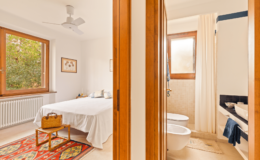 Bright and airy bedroom featuring a white bedspread, wooden furniture, and large windows with natural light. Adjacent to the bedroom is a modern bathroom with a shower and two toilets, separated by a wooden door.