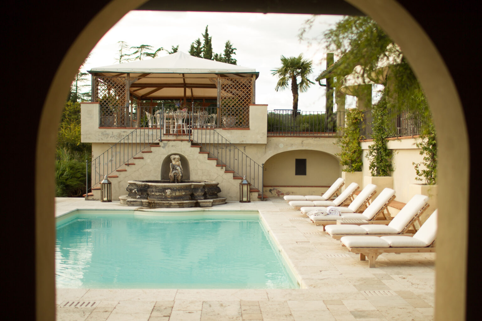 View of a serene swimming pool surrounded by lounge chairs, with a decorative fountain and a gazebo on a terrace in the background, set in a lush garden landscape. Perfect for relaxation and outdoor gatherings. | Only on SopranoVillas