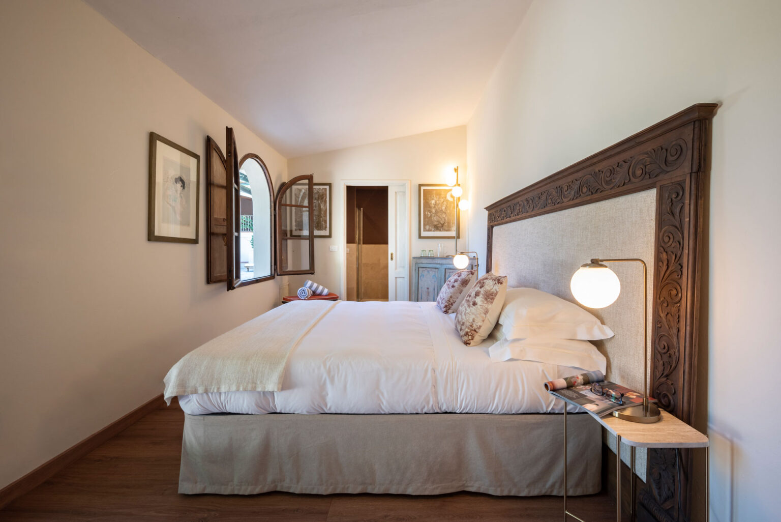 Cozy bedroom featuring a large bed with decorative pillows, rustic wooden headboard, and bedside lamp. The room has large open windows with wooden shutters, allowing natural light to fill the space. Tasteful artwork adorns the walls, complemented by a stylish side table displaying magazines, creating a warm and inviting atmosphere. | Only on SopranoVillas