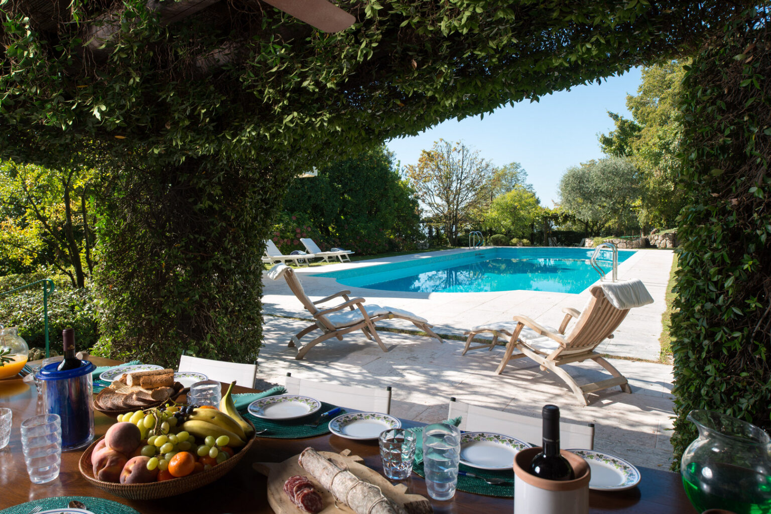 Outdoor dining area with a view of a swimming pool surrounded by lush greenery, featuring a table set with fruit, bread, and drinks, and loungers positioned by the poolside. Perfect for relaxation and entertaining in a serene environment. | Only on SopranoVillas