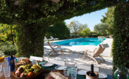 Outdoor dining area with a view of a swimming pool surrounded by lush greenery, featuring a table set with fruit, bread, and drinks, and loungers positioned by the poolside. Perfect for relaxation and entertaining in a serene environment.