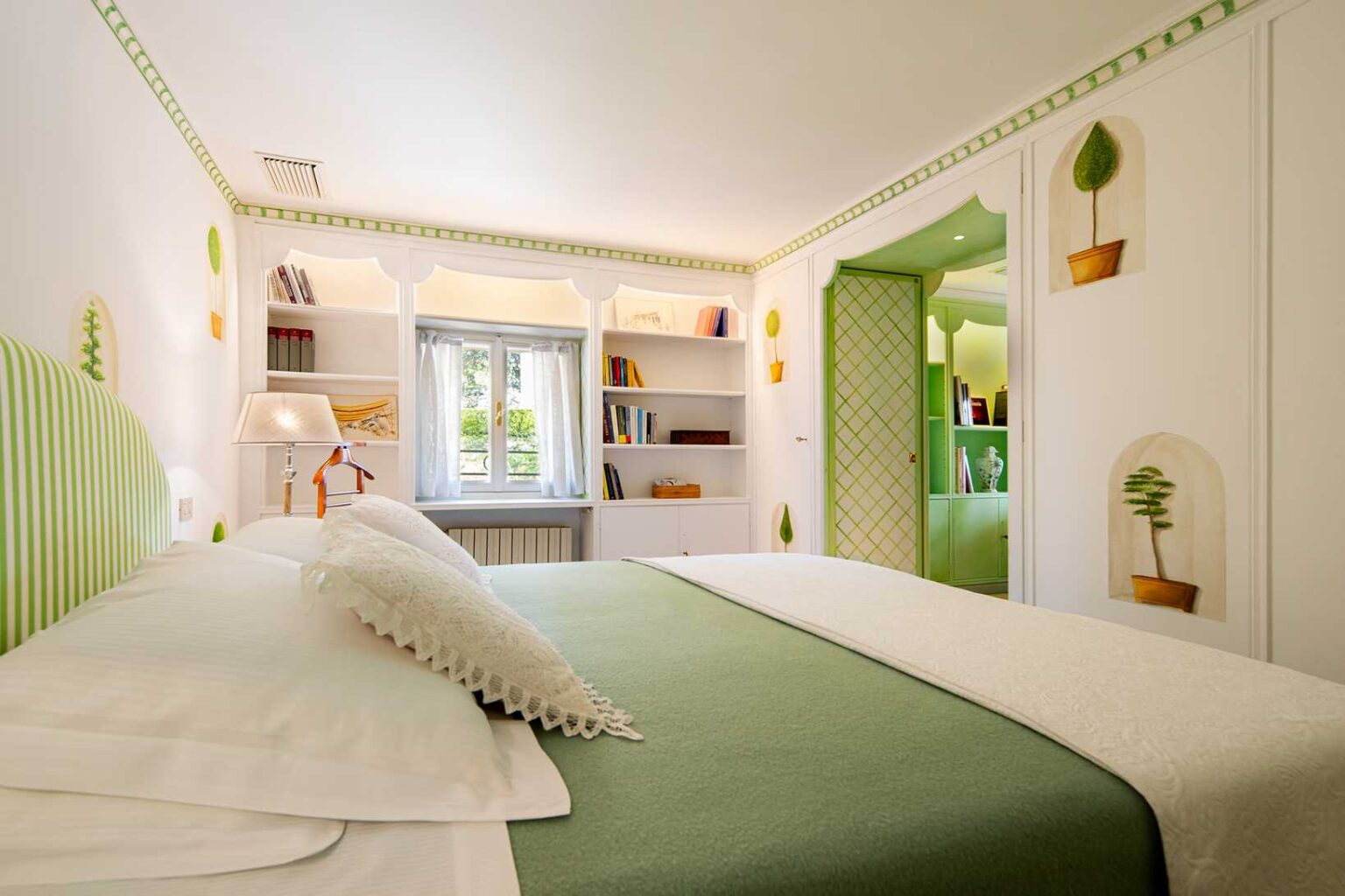 Bright and stylish bedroom featuring a green striped headboard, comfortable bedding, and decorative wall art. The room includes a window with sheer curtains, a bedside lamp, and a bookshelf filled with books, creating a cozy and inviting atmosphere. | Only on SopranoVillas