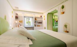 Bright and stylish bedroom featuring a green striped headboard, comfortable bedding, and decorative wall art. The room includes a window with sheer curtains, a bedside lamp, and a bookshelf filled with books, creating a cozy and inviting atmosphere.