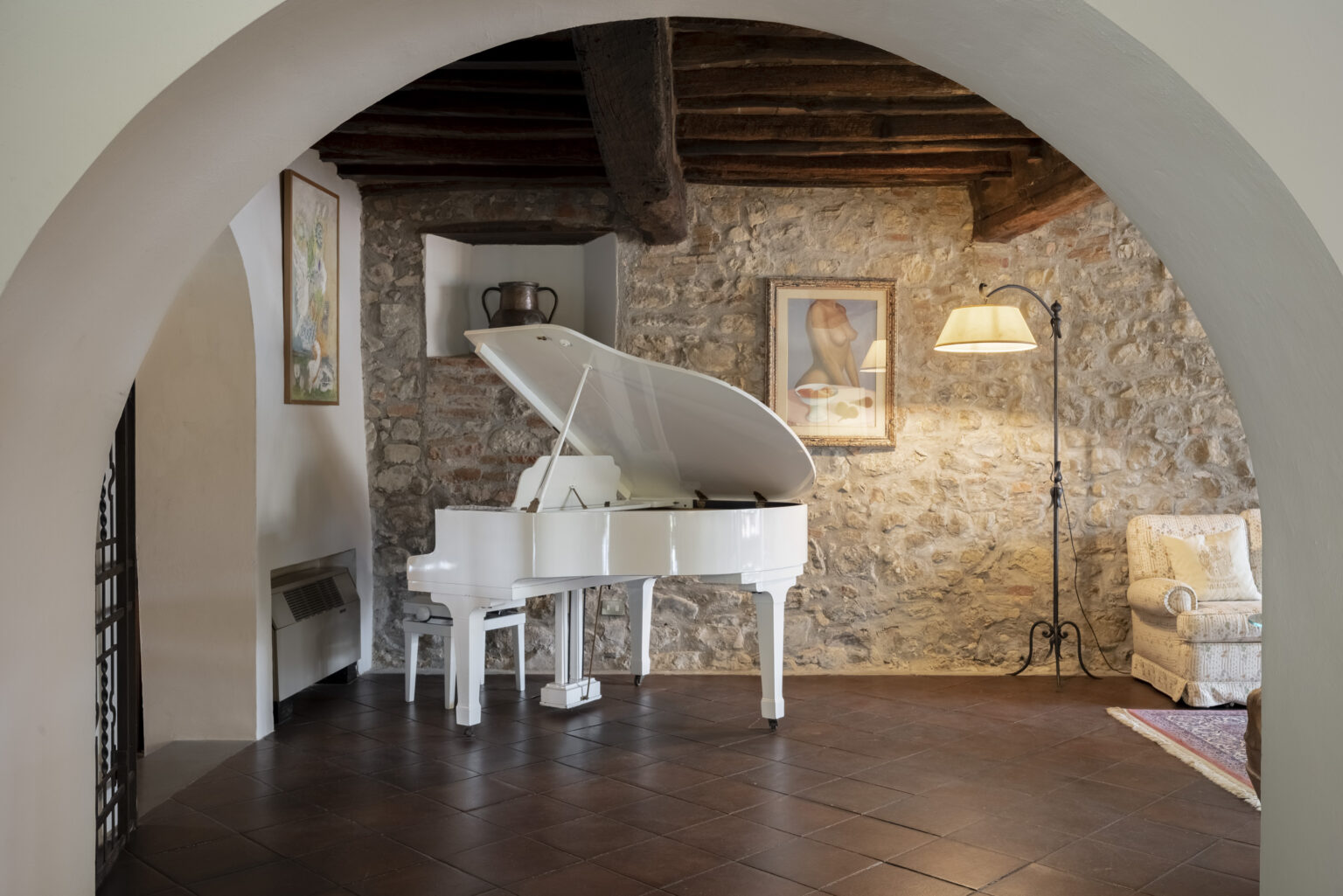 Elegant white grand piano in a cozy living room with stone walls, framed artwork, and a comfortable armchair, illuminated by a stylish floor lamp. | Only on SopranoVillas