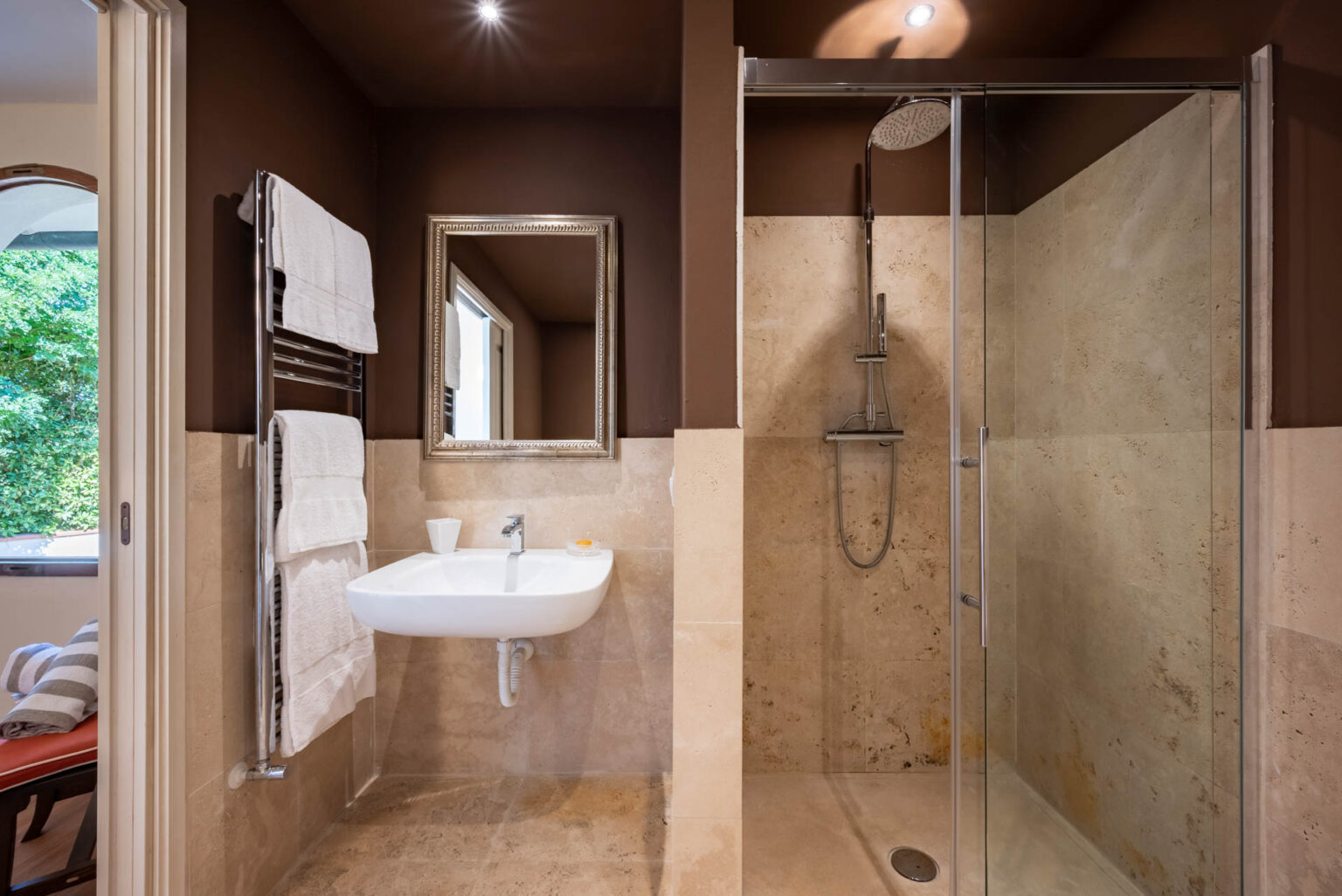Modern bathroom featuring a walk-in shower with glass enclosure, a sleek wall-mounted sink, and a towel rack with neatly arranged towels. The walls are painted in a rich brown color, complemented by natural stone tiles on the floor and shower area. A decorative mirror adds a touch of elegance, while natural light filters in from a window. | Only on SopranoVillas