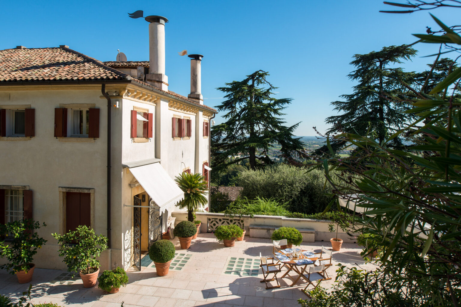 Charming Italian villa with a terracotta roof, featuring large windows with wooden shutters, surrounded by lush greenery and potted plants. A sunlit terrace showcases a dining table set for outdoor meals, with a scenic view of the countryside in the background. | Only on SopranoVillas