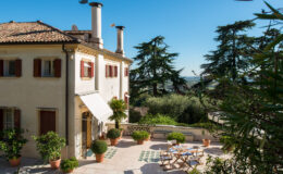Charming Italian villa with a terracotta roof, featuring large windows with wooden shutters, surrounded by lush greenery and potted plants. A sunlit terrace showcases a dining table set for outdoor meals, with a scenic view of the countryside in the background.