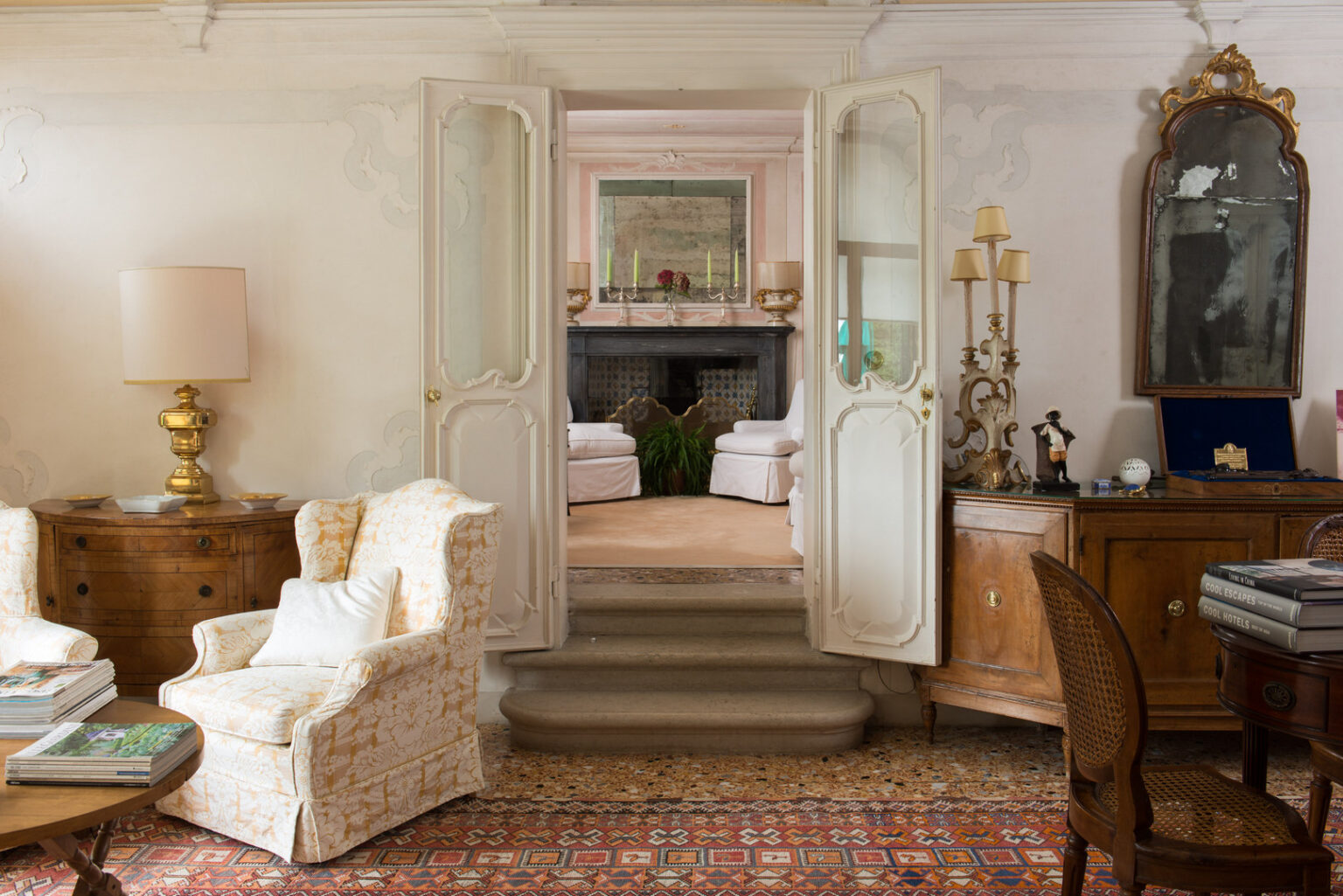Elegant interior of a classic living room featuring floral-patterned armchairs, a wooden coffee table with magazines, and a decorative rug. The room opens to a cozy area beyond double doors, showcasing a fireplace, plants, and soft furnishings. Antique furniture and lamps add a touch of vintage charm to the space. | Only on SopranoVillas