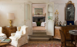 Elegant interior of a classic living room featuring floral-patterned armchairs, a wooden coffee table with magazines, and a decorative rug. The room opens to a cozy area beyond double doors, showcasing a fireplace, plants, and soft furnishings. Antique furniture and lamps add a touch of vintage charm to the space.
