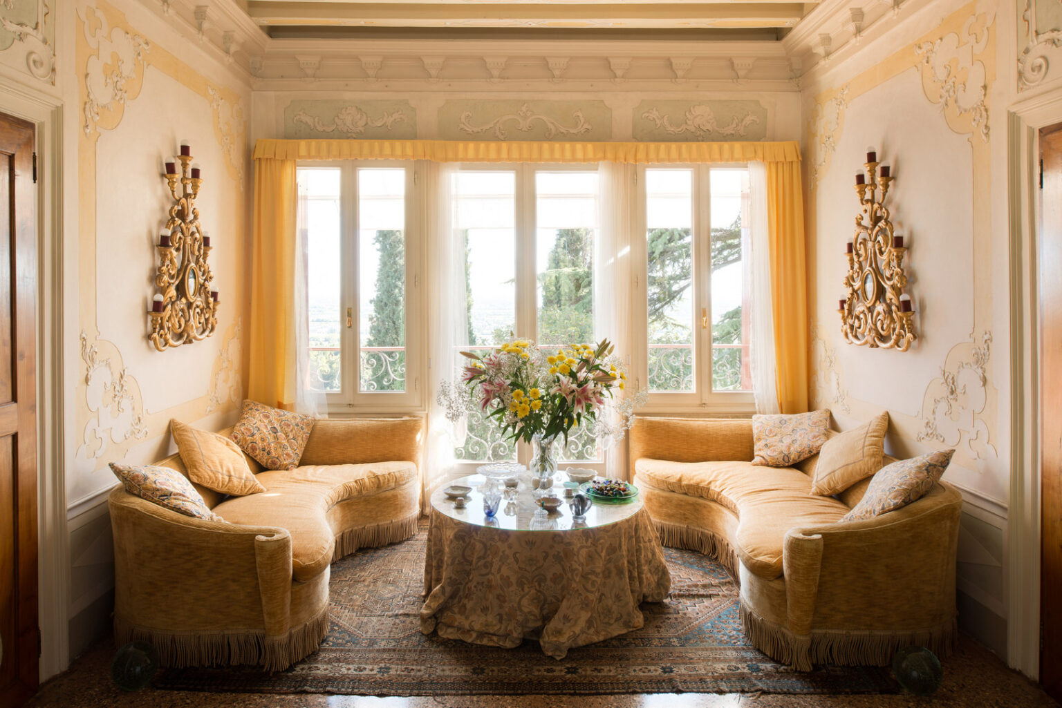 Elegant living room featuring two curved, yellow velvet sofas adorned with decorative pillows, a round glass-top table draped with a floral tablecloth, and a vibrant bouquet of flowers. Natural light streams through large windows, highlighting ornate wall decorations and a vintage wall sconce. The space exudes a warm and inviting atmosphere. | Only on SopranoVillas