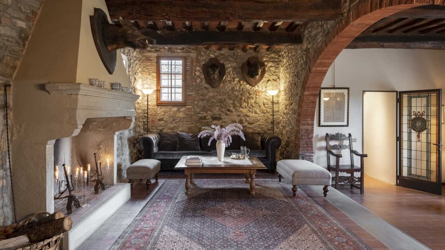 Cozy living room featuring a stone wall, wooden beams, and a decorative fireplace. The space includes a dark leather sofa, two upholstered benches, a wooden coffee table with a vase of flowers, and ambient lighting. A vintage rug adds warmth, while a decorative chair and an open door lead to another room. | Only on SopranoVillas