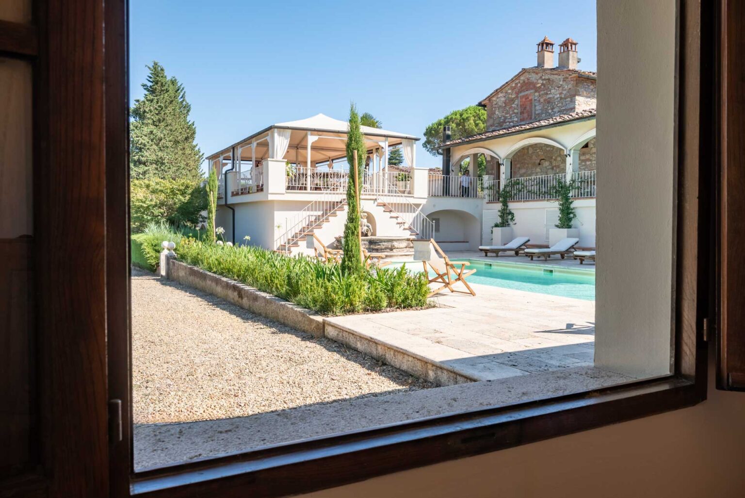 View of a luxurious villa with a swimming pool, surrounded by lush greenery and a gravel pathway, as seen through a window. The villa features a spacious terrace with seating and a staircase leading to the upper level, showcasing a charming architectural design under a clear blue sky. | Only on SopranoVillas