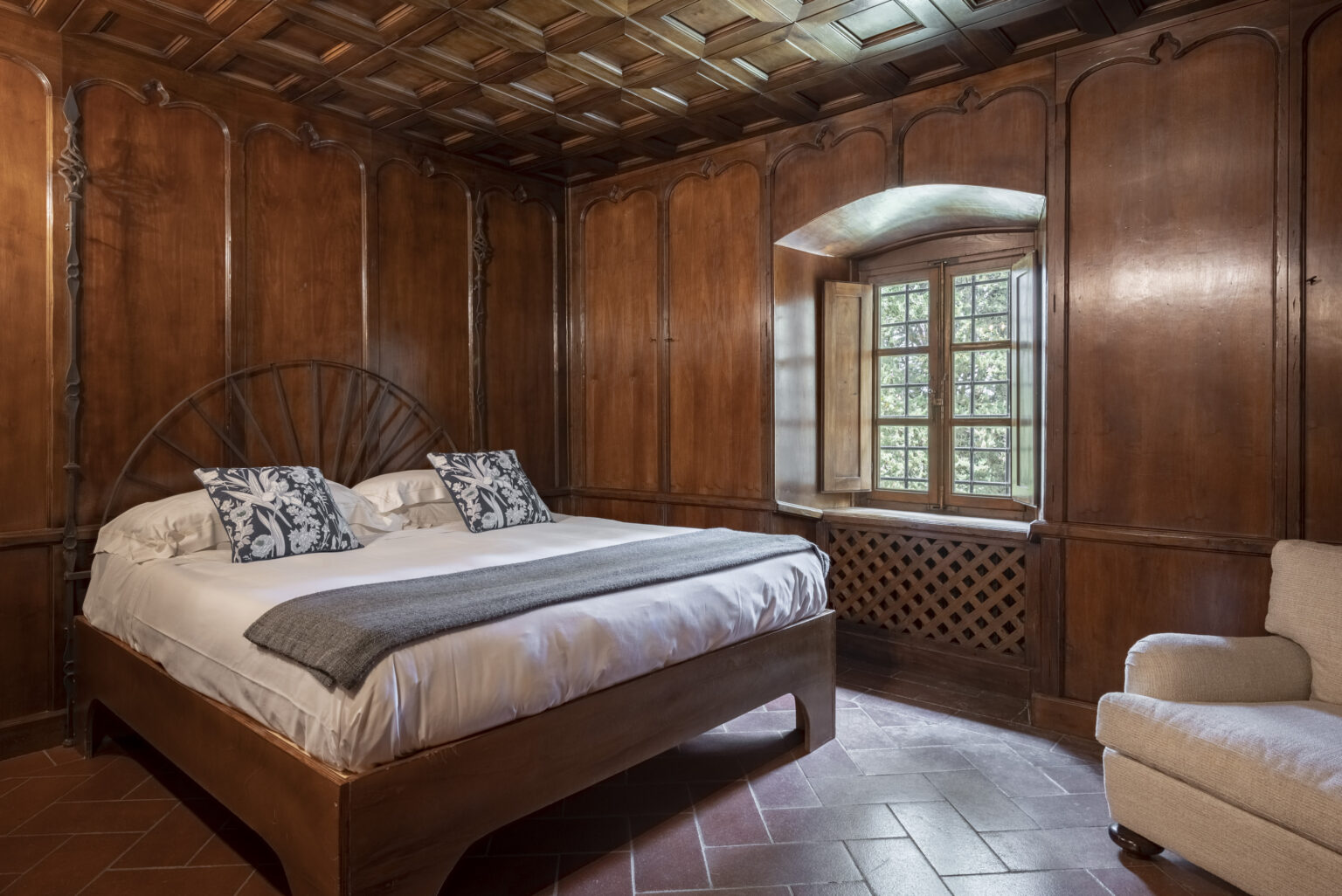 Cozy bedroom featuring a wooden bed with decorative pillows, a plush gray throw, and rich wooden paneling. The room includes a window with a view of greenery, allowing natural light to enhance the warm ambiance. Ideal for a relaxing retreat or a romantic getaway. | Only on SopranoVillas