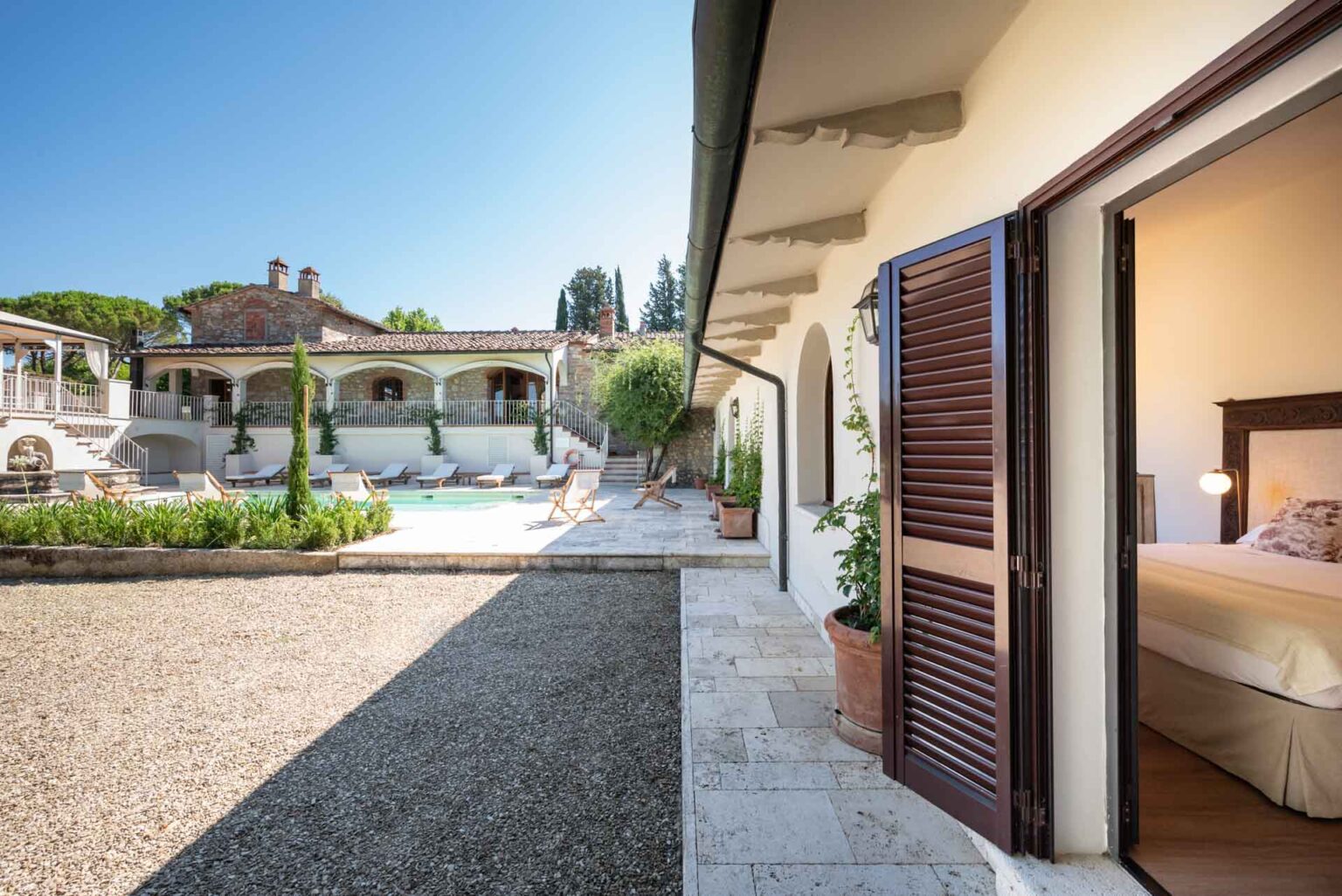 Elegant villa exterior with a view of a swimming pool, lounge chairs, and lush greenery. The image shows a doorway leading into a cozy bedroom, highlighting the serene atmosphere of the property. Perfect for luxury getaways in a picturesque setting. | Only on SopranoVillas