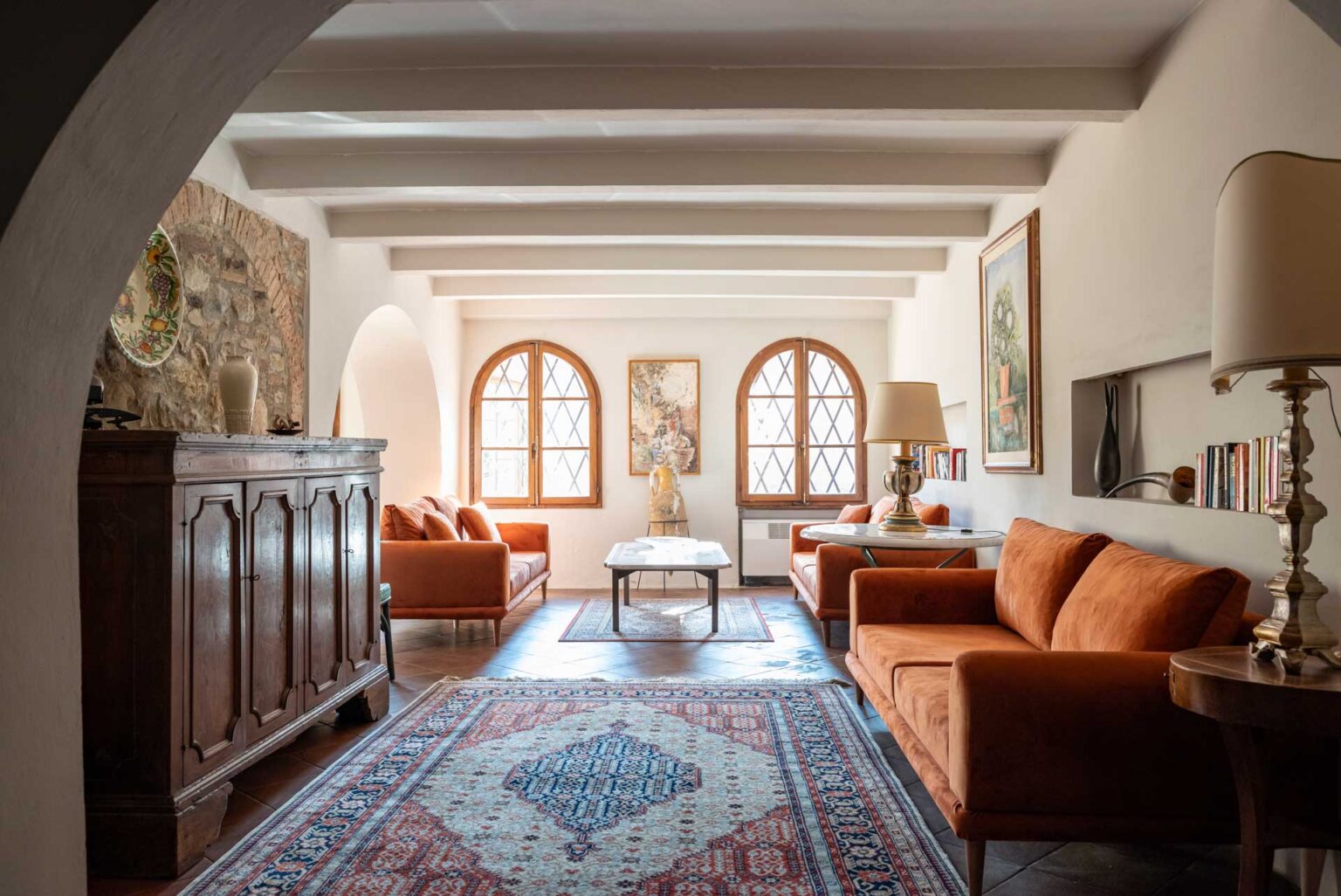 Living room featuring warm orange sofas, a wooden cabinet, and decorative art pieces. Natural light streams through arched windows, illuminating a patterned area rug and a stylish coffee table. The space combines rustic charm with modern decor elements, creating a cozy ambiance. | Only on SopranoVillas
