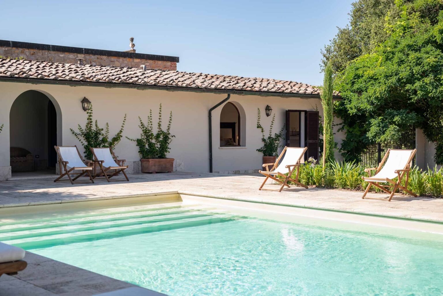 Serene outdoor patio featuring a sparkling swimming pool, two wooden lounge chairs, and a charming villa with a tiled roof and lush greenery, perfect for relaxation and leisure. | Only on SopranoVillas