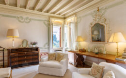 Elegant living room featuring ornate wall decor, wooden furniture, and soft lighting. The space includes a vintage dresser, a decorative mirror, and comfortable seating adorned with floral cushions. Large windows invite natural light and showcase a scenic view, enhancing the room's inviting atmosphere.
