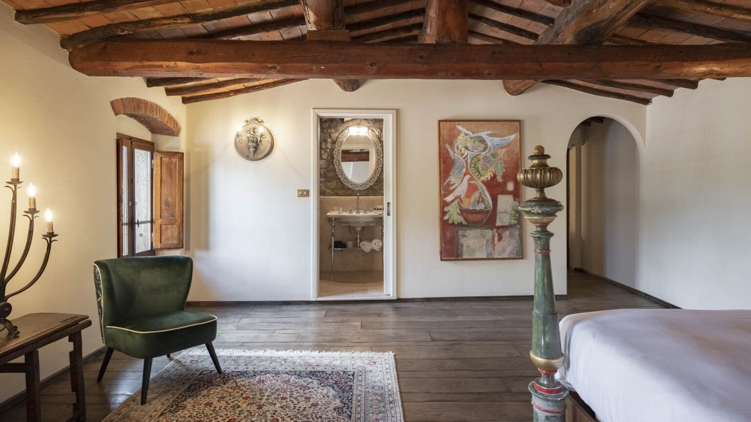 Cozy bedroom with rustic wooden beams, featuring a green accent chair, a decorative rug, and an artistic painting on the wall. The room includes a doorway leading to a stylish bathroom with vintage decor. Natural light floods in through the open window, enhancing the warm atmosphere. | Only on SopranoVillas