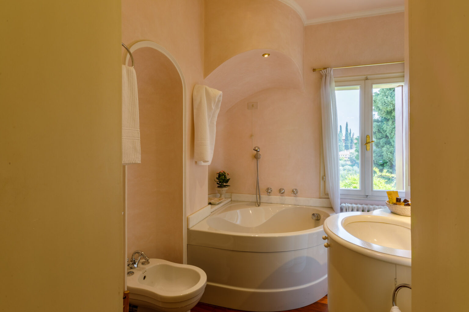 Modern bathroom featuring a curved bathtub, bidet, and elegant decor with a soft pink color scheme. Natural light filters through a window, illuminating the space and highlighting the stylish fixtures and towels. | Only on SopranoVillas