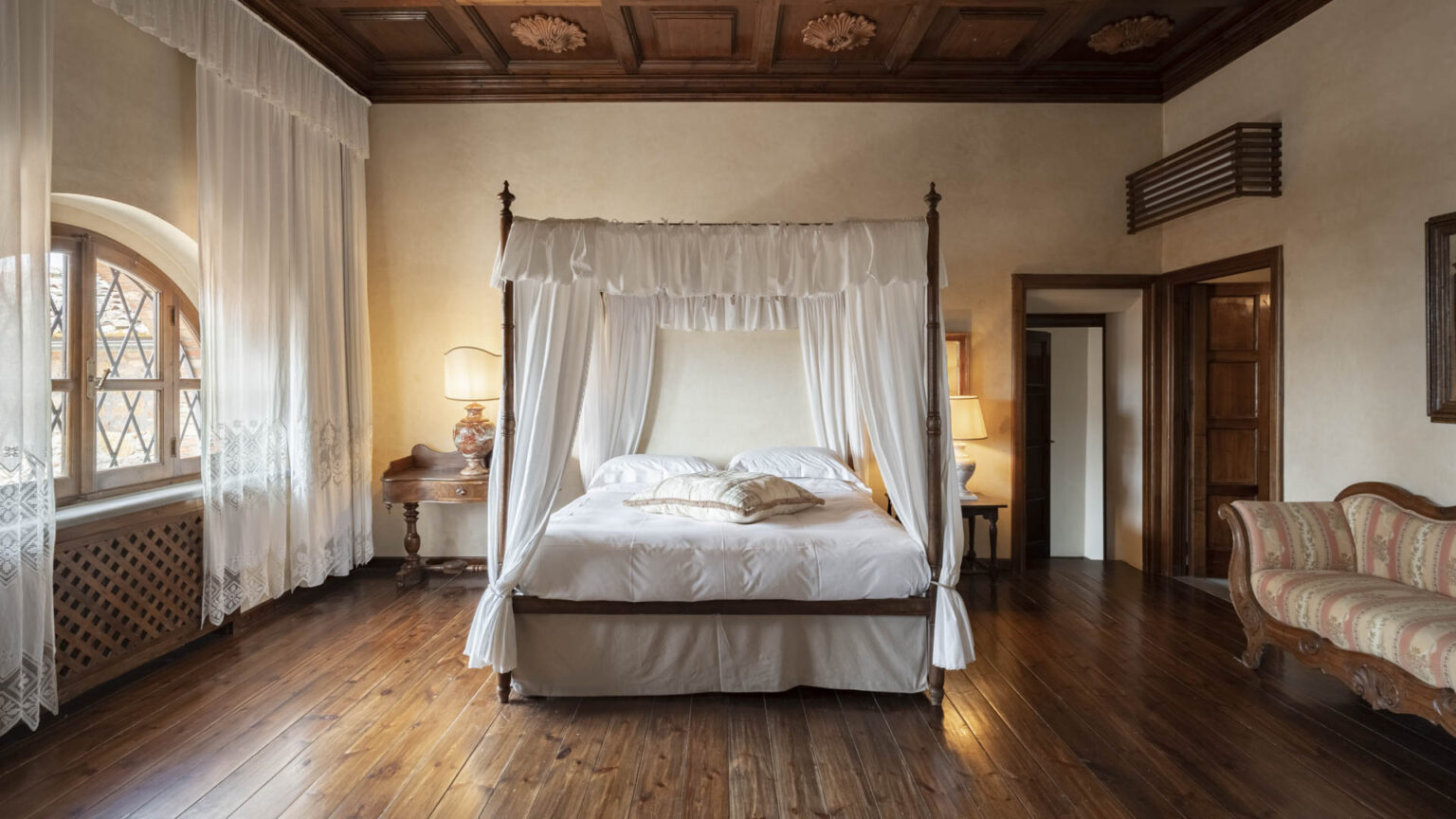 Elegant bedroom featuring a four-poster bed with white drapes, hardwood flooring, and natural light from arched windows. The room includes a vintage bedside table with a lamp and a classic upholstered sofa, creating a cozy and inviting atmosphere. Ideal for relaxation and comfort. | Only on SopranoVillas