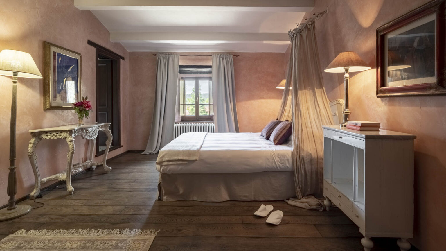 Cozy bedroom interior featuring soft pink walls, a comfortable double bed with decorative pillows, elegant lighting, and vintage furniture. Natural light streams through a window adorned with light curtains, creating a warm and inviting atmosphere. | Only on SopranoVillas
