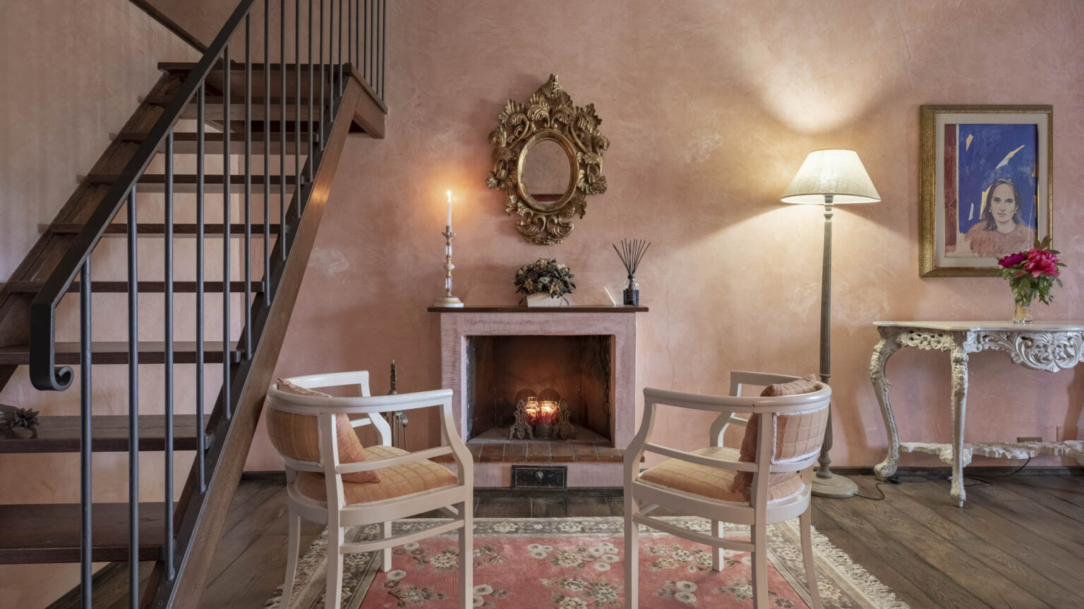 Cozy living room featuring a pink plaster wall, a white fireplace with candles, two elegant chairs, a decorative mirror, and a stylish lamp, complemented by a floral arrangement on a vintage side table. | Only on SopranoVillas