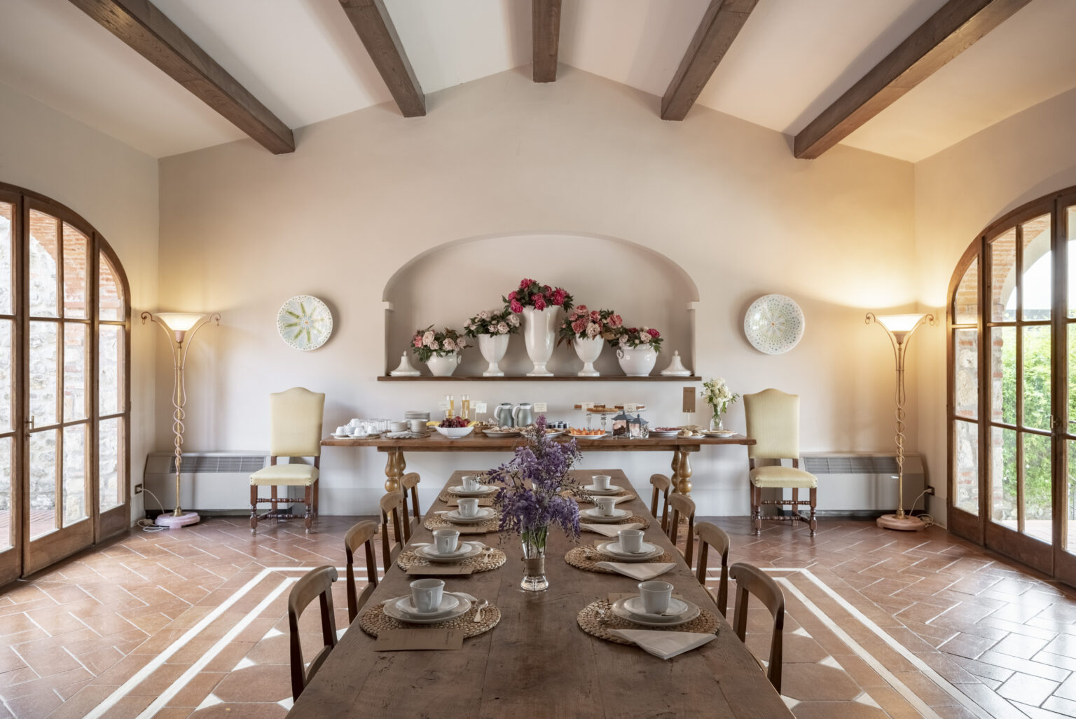 Elegant dining room featuring a long wooden table set for breakfast, adorned with white plates and woven placemats. The room showcases a floral centerpiece, large windows, and decorative lighting, creating a warm and inviting atmosphere. A side table displays a variety of breakfast items, enhancing the cozy and welcoming ambiance. | Only on SopranoVillas