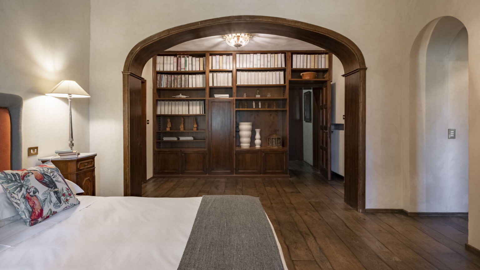 Cozy bedroom featuring a stylish bed with a decorative pillow, a bedside lamp, and a wooden nightstand. The room has a warm ambiance with wooden flooring and an arched doorway leading to a bookshelf filled with books and decorative items. Natural light illuminates the space, enhancing its inviting atmosphere. | Only on SopranoVillas
