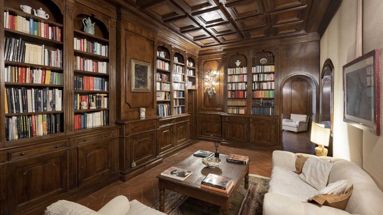 Cozy library interior featuring dark wooden shelves filled with books, a wooden coffee table with magazines, and a comfortable white sofa. Elegant wall sconces provide warm lighting, enhancing the inviting atmosphere of the reading space. | Only on SopranoVillas