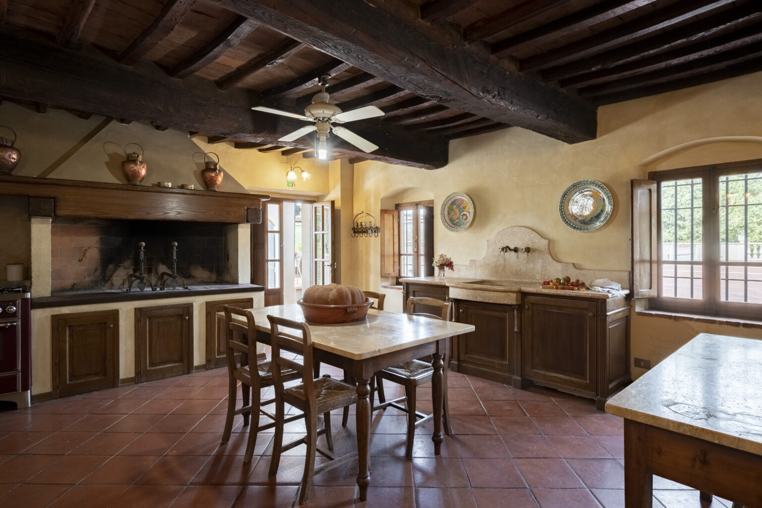 Rustic kitchen interior featuring wooden beams, a large central dining table, and traditional decor. The space includes a stone fireplace, wooden cabinetry, and natural light streaming through windows, creating a warm and inviting atmosphere. Ideal for cozy family meals and gatherings. | Only on SopranoVillas