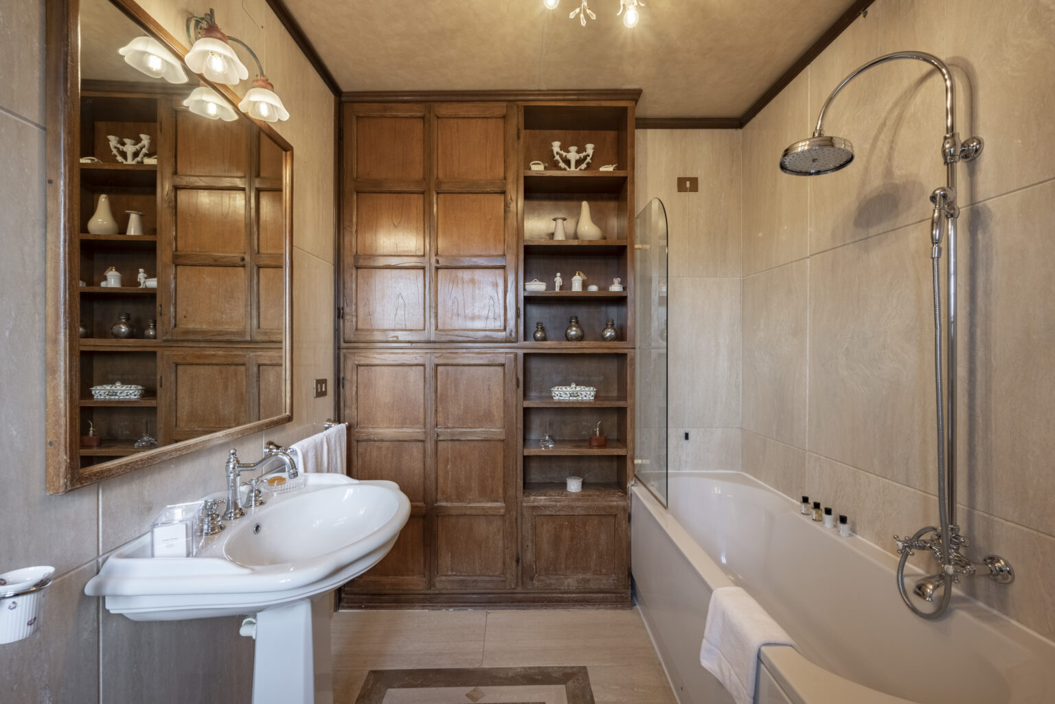 Spacious and elegant bathroom featuring a white sink with vintage fixtures, a wooden cabinet with decorative items, a glass shower, and a bathtub. Soft lighting enhances the warm tones of the wooden elements and tile finishes. | Only on SopranoVillas
