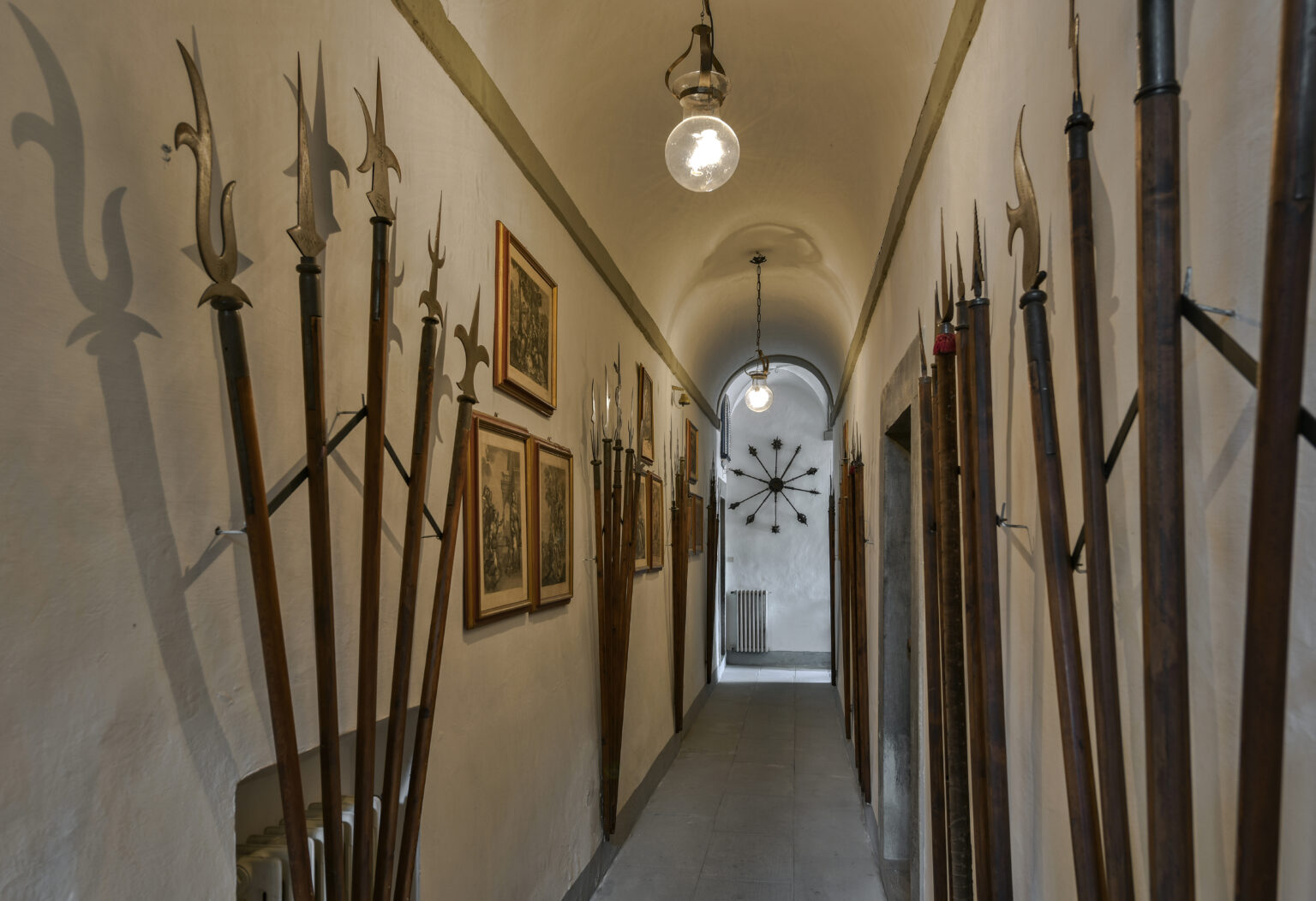 Historic hallway featuring ornate spears mounted on the walls, framed artwork depicting historical scenes, and vintage lighting fixtures, creating an atmosphere of antiquity and craftsmanship. | Only on SopranoVillas