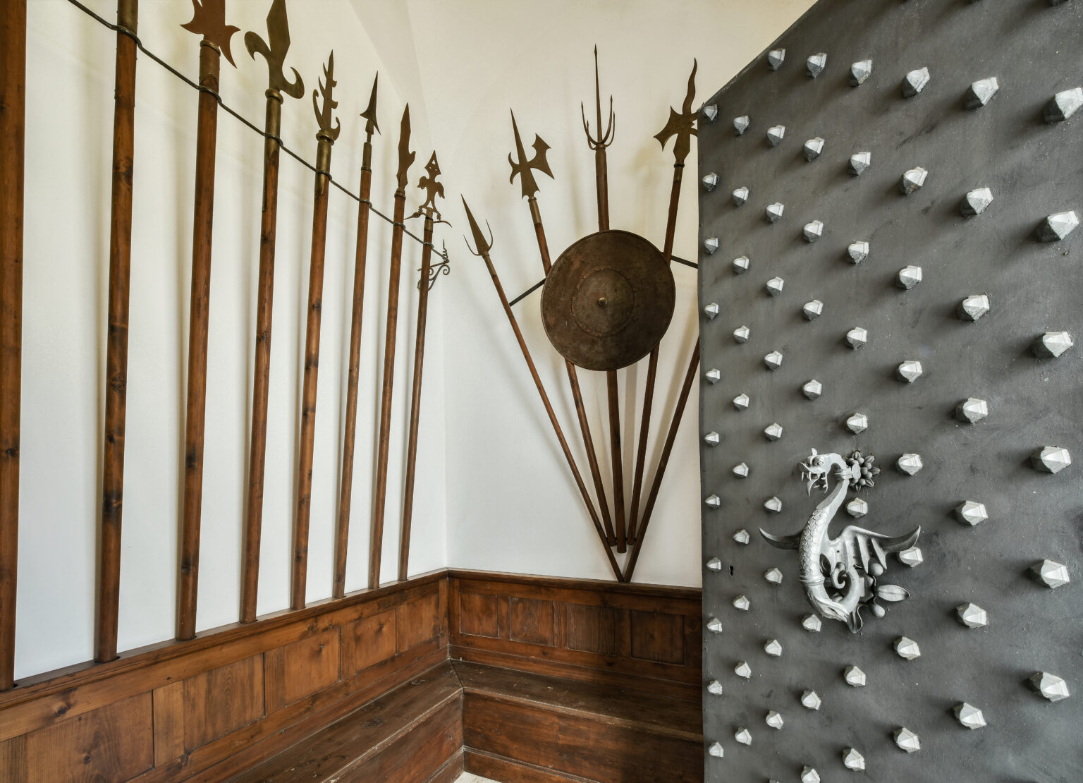 Historic weapons display featuring ornate spears and a shield mounted on a wooden wall, alongside a textured door adorned with decorative spikes and a dragon emblem. | Only on SopranoVillas